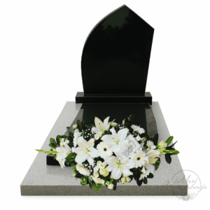 Fekete gránit síremlék előtt elhelyezett kegyeleti sírpárna fehér liliomokkal, rózsákkal és krizantémokkal, zöld lombbal és „RIP” szalaggal, a Mesi Virágoskertje kompozíciója.