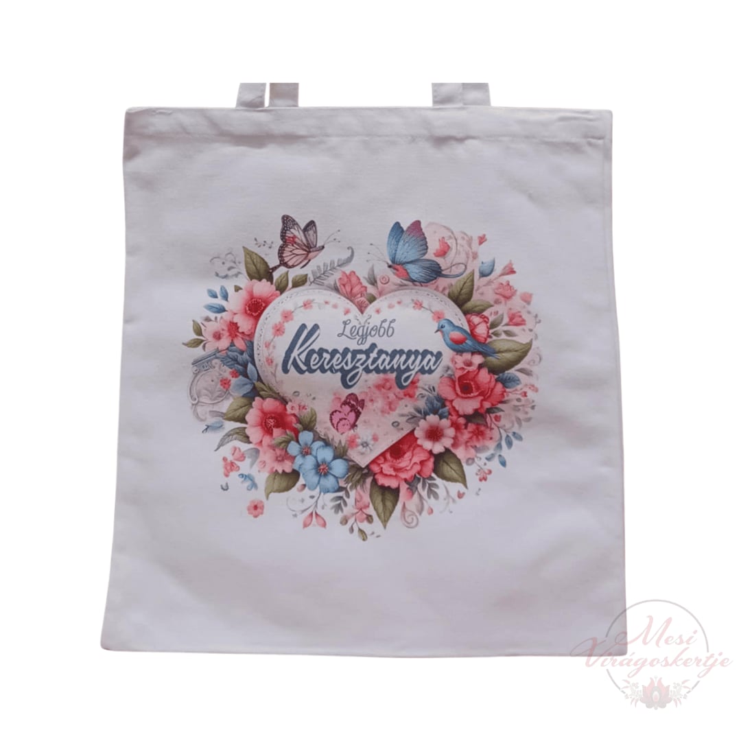 Fehér vászontáska, amelyen egy szív alakú minta látható, körülötte virágokkal, pillangókkal és madarakkal. A szív közepén a „Legjobb Keresztanya” felirat olvasható.