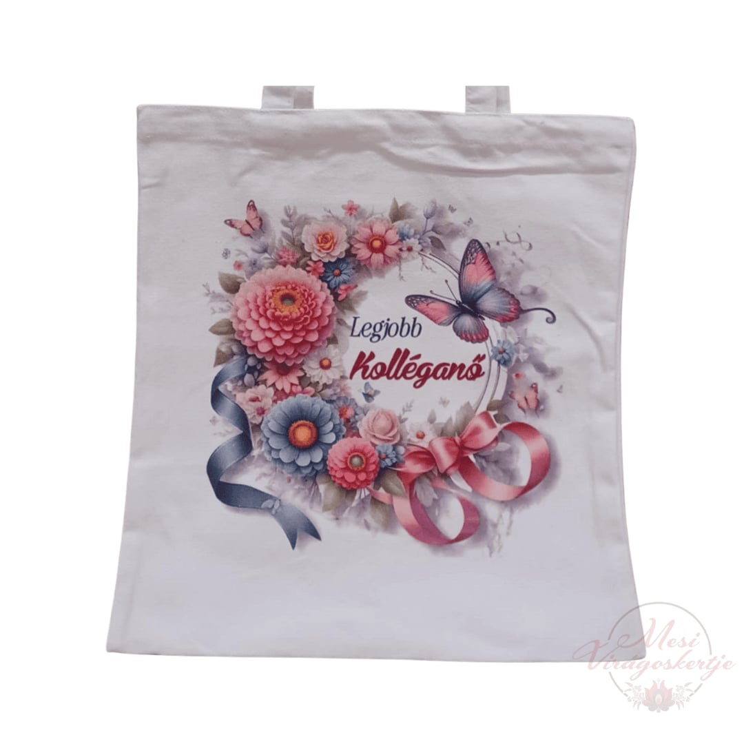 Fehér vászontáska, rajta színes virágkoszorú, közepén rózsaszín-lila pillangóval és „Legjobb kolléganő” felirattal. A koszorút rózsaszín és kék szalagok díszítik, jobb alsó sarokban „Mesi Virágoskertje” logóval.
