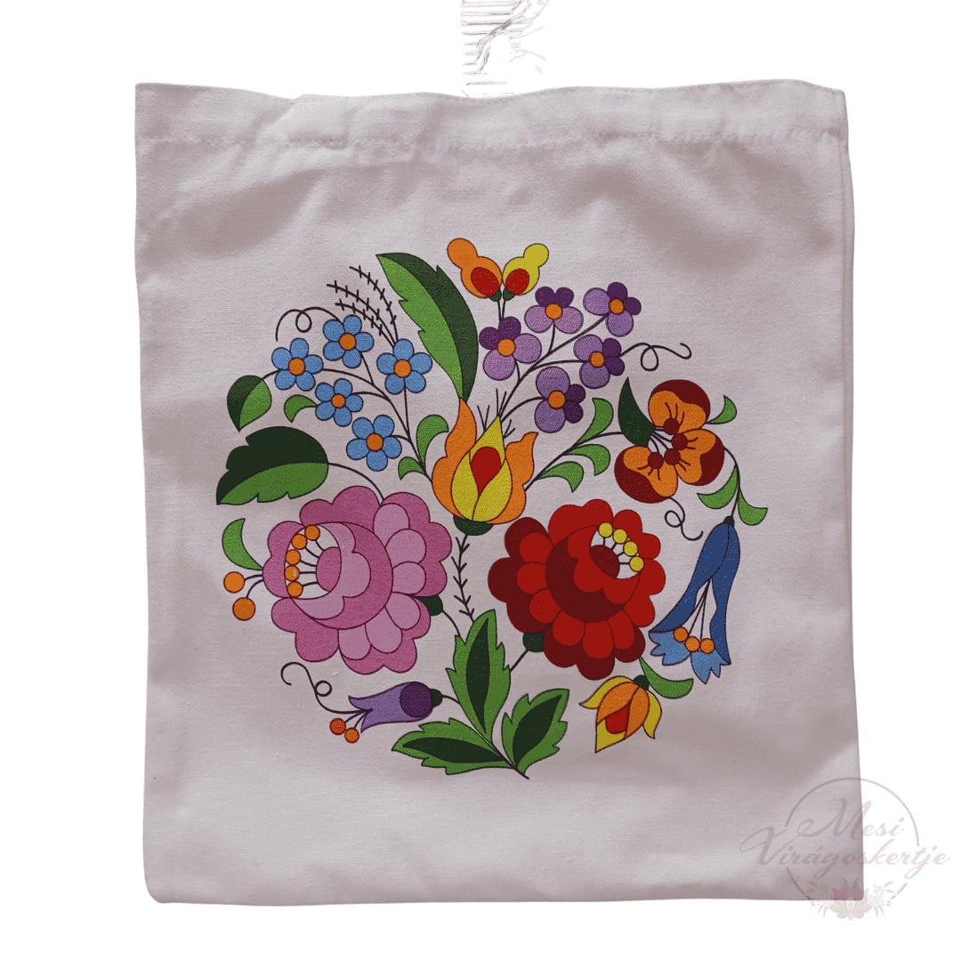 Fehér vászontáska színes virágkoszorúval, amely piros, rózsaszín, sárga, narancssárga, lila és kék virágokból áll, zöld levelekkel kiegészítve. A táska jobb alsó sarkában „Mesi Virágoskertje” vízjel látható.